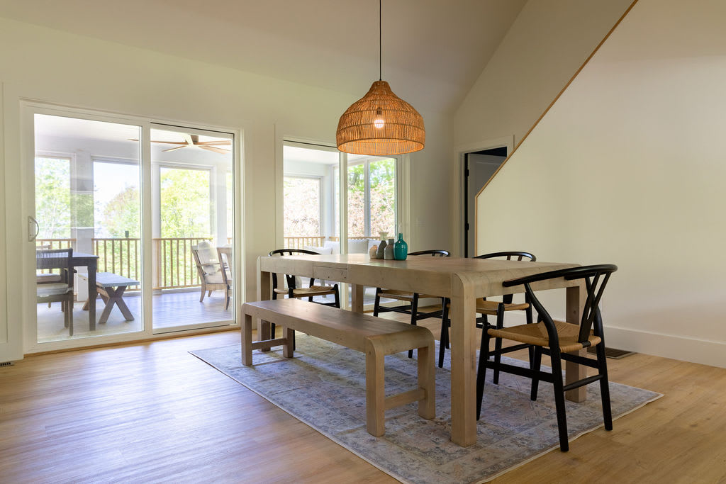 A bright dining area featuring a large light wood dining table with a bench and several black and woven chairs, set on an area rug. Sliding glass doors lead to an outdoor porch, and a large woven pendant light hangs above the table.