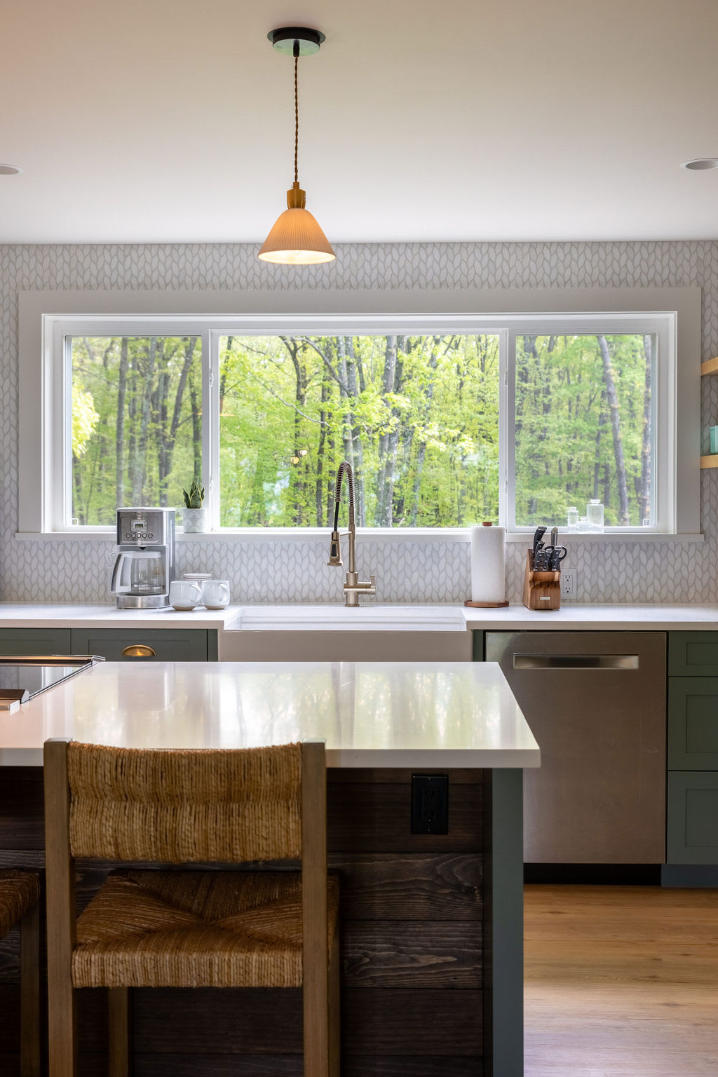 Close-up of the kitchen island with white quartz countertops, a farmhouse sink, and a large window offering expansive views of the surrounding forest. The backsplash features a subtle patterned tile.