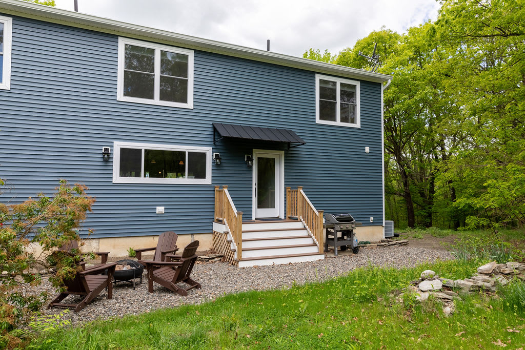 The side of the blue house features a white door with a black awning, leading to wooden stairs, and a gravel patio with Adirondack chairs and a fire pit, surrounded by green grass and trees.