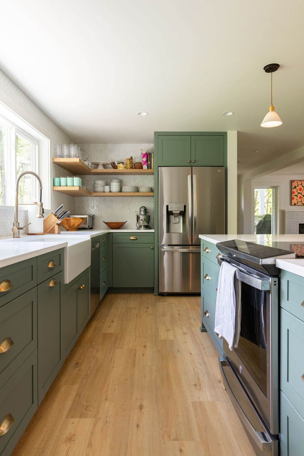 A modern kitchen features sage green cabinetry with brass hardware, a white farmhouse sink, and stainless steel appliances including a refrigerator and range. Open wooden shelves display dishware, and light wood flooring runs throughout the space.