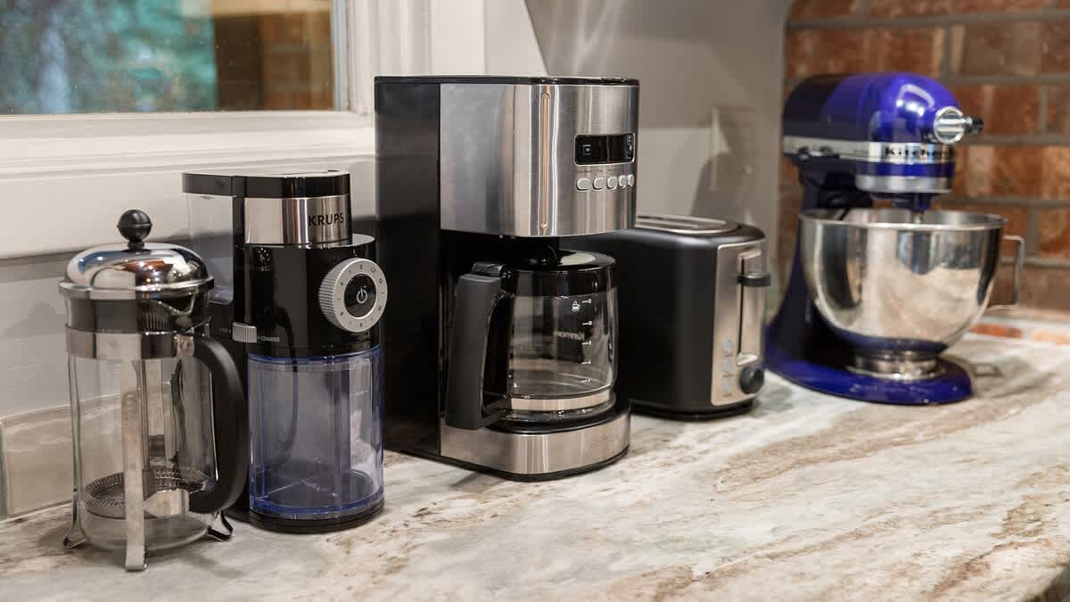 Close-up of a kitchen countertop showcasing various small appliances, including a coffee maker, coffee grinder, toaster, and stand mixer, set on a light marble-look surface.