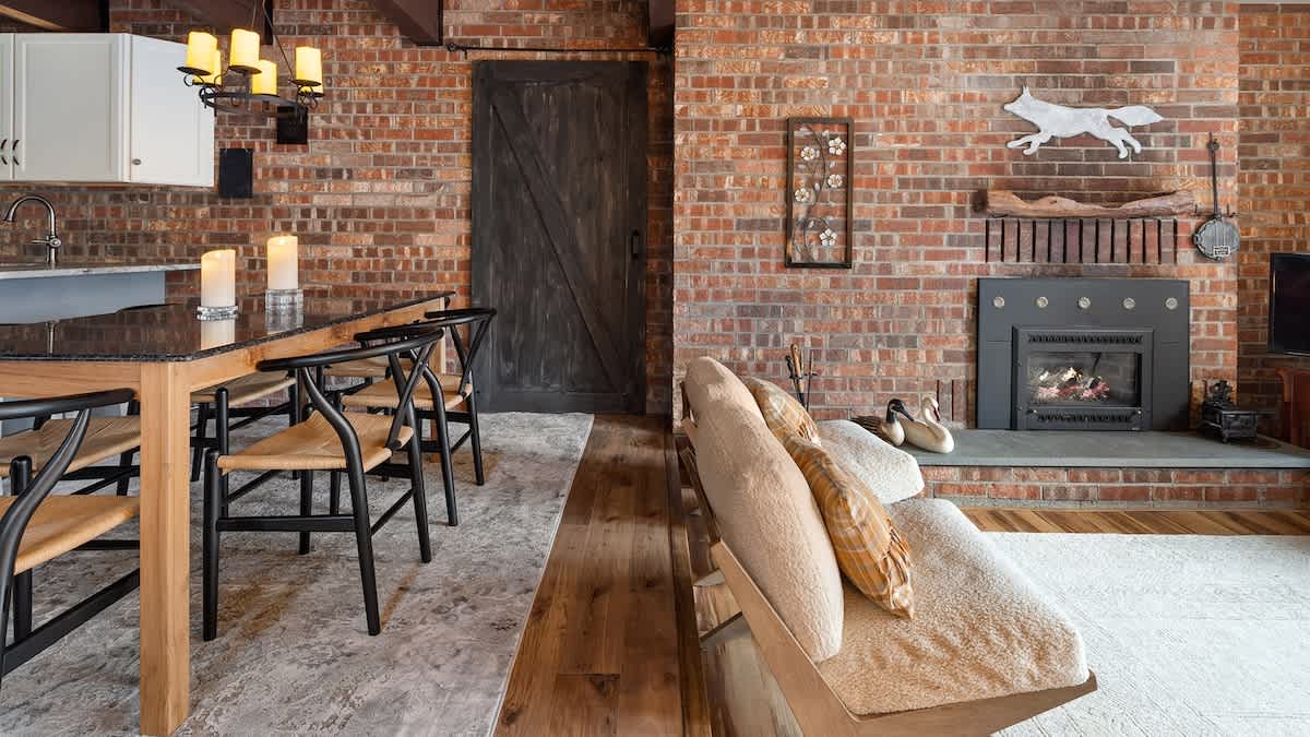 Inviting living room with a brick fireplace and a white fox wall decoration, adjacent to a dining area with a dark granite table and black chairs, all on light hardwood floors.