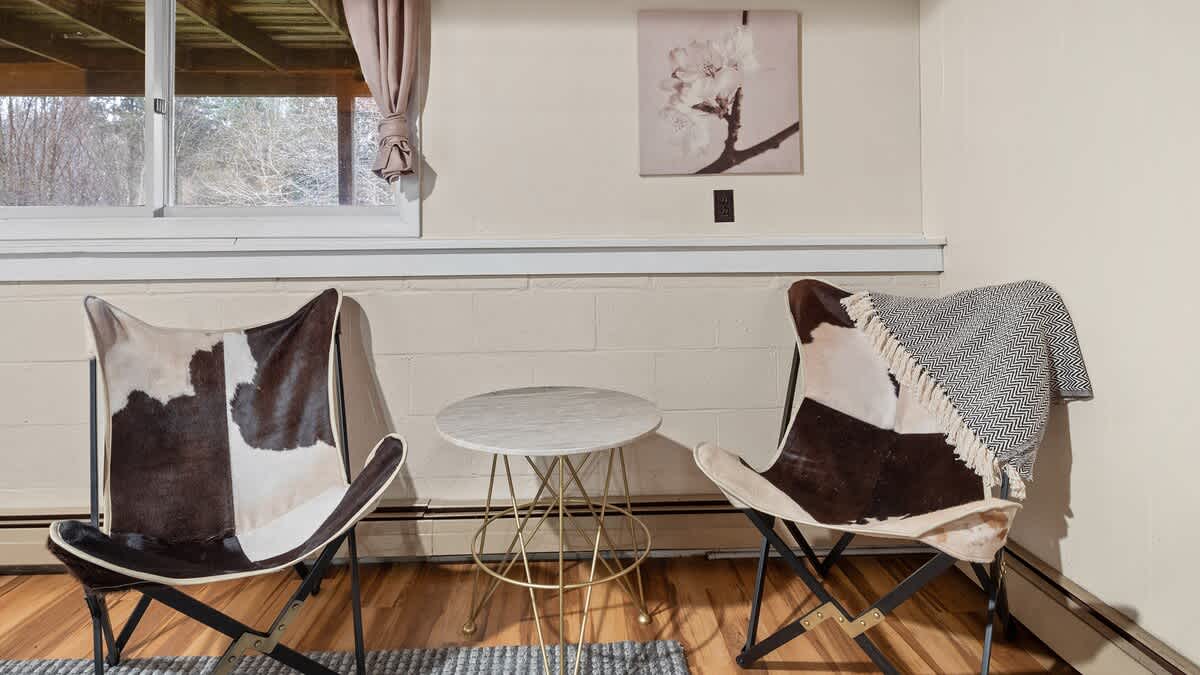 A cozy sitting area features two faux cowhide butterfly chairs flanking a small round marble-top table, positioned beneath a window with a view of trees. A patterned throw blanket is draped over one chair.