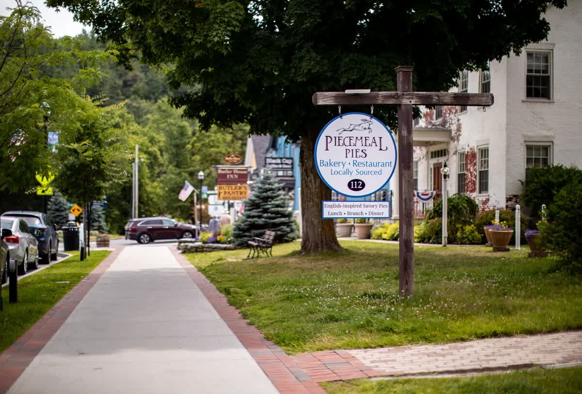 A quaint street scene features a prominent wooden sign for "Piecemeal Pies Bakery - Restaurant" on a grassy lawn, with a sidewalk, parked cars, and a white building in the background. The image captures a charming local business.