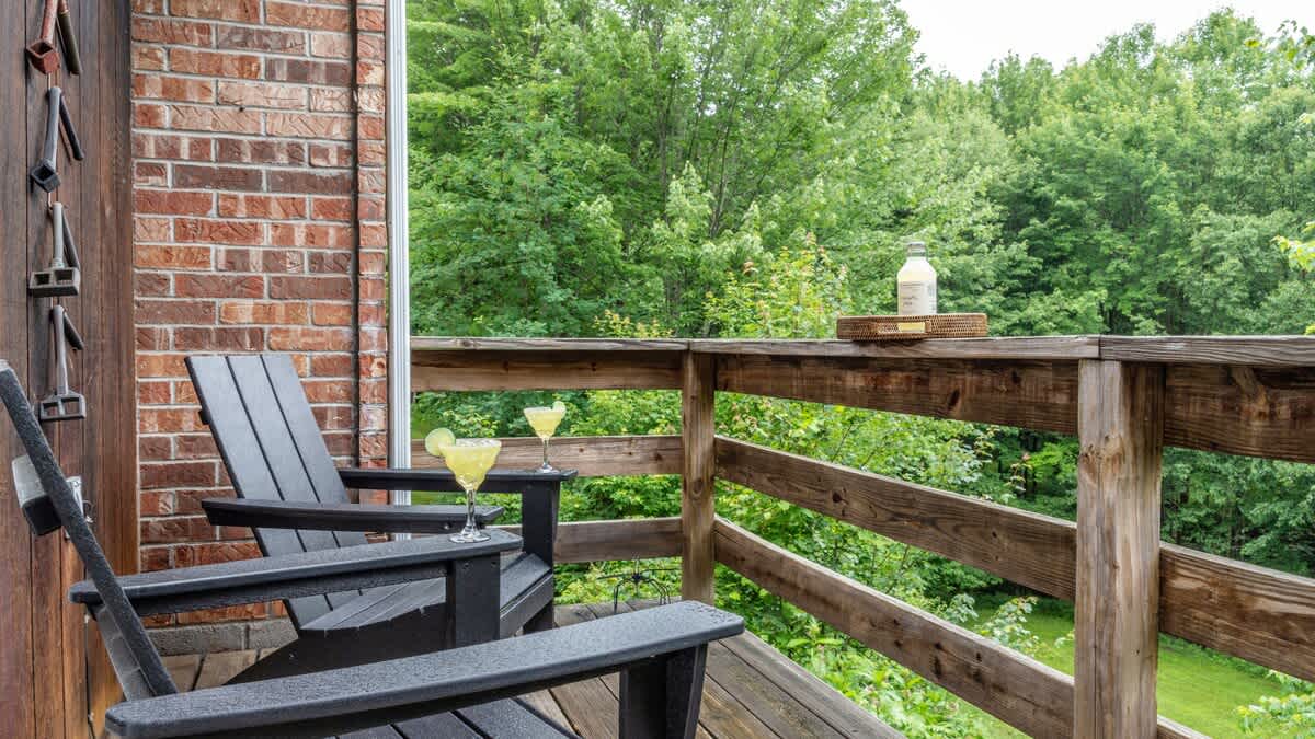 Two dark Adirondack chairs are placed on a wooden deck, with two cocktails on a small table between them, offering a serene view of the dense green forest through the wooden railing.