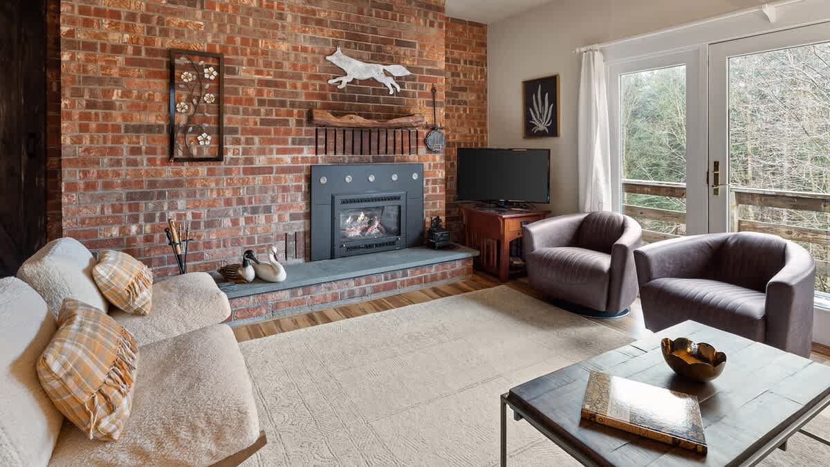 A spacious living room featuring a brick accent wall with a fireplace, a light-colored sofa, two upholstered armchairs, a flat-screen TV, and French doors leading to an outdoor area.