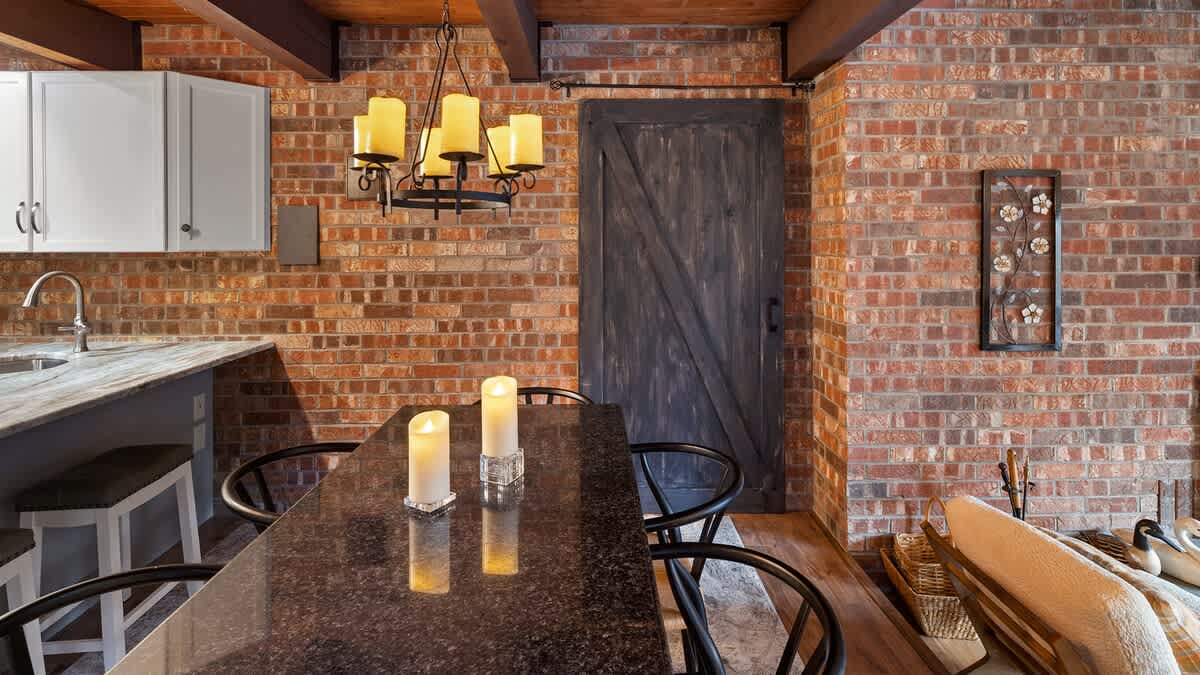 Dining area featuring a dark granite-top table with lit candles and black chairs, set against a rustic brick wall with a dark wooden barn door, and a modern kitchen visible in the background.