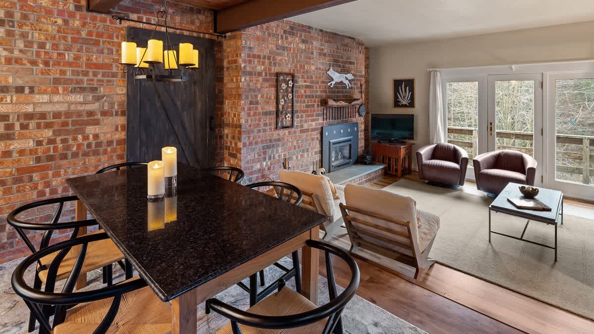 A dining area with a dark granite-topped table and black wooden chairs, illuminated by a chandelier with candle-style lights, adjacent to a brick fireplace and a living area with armchairs.