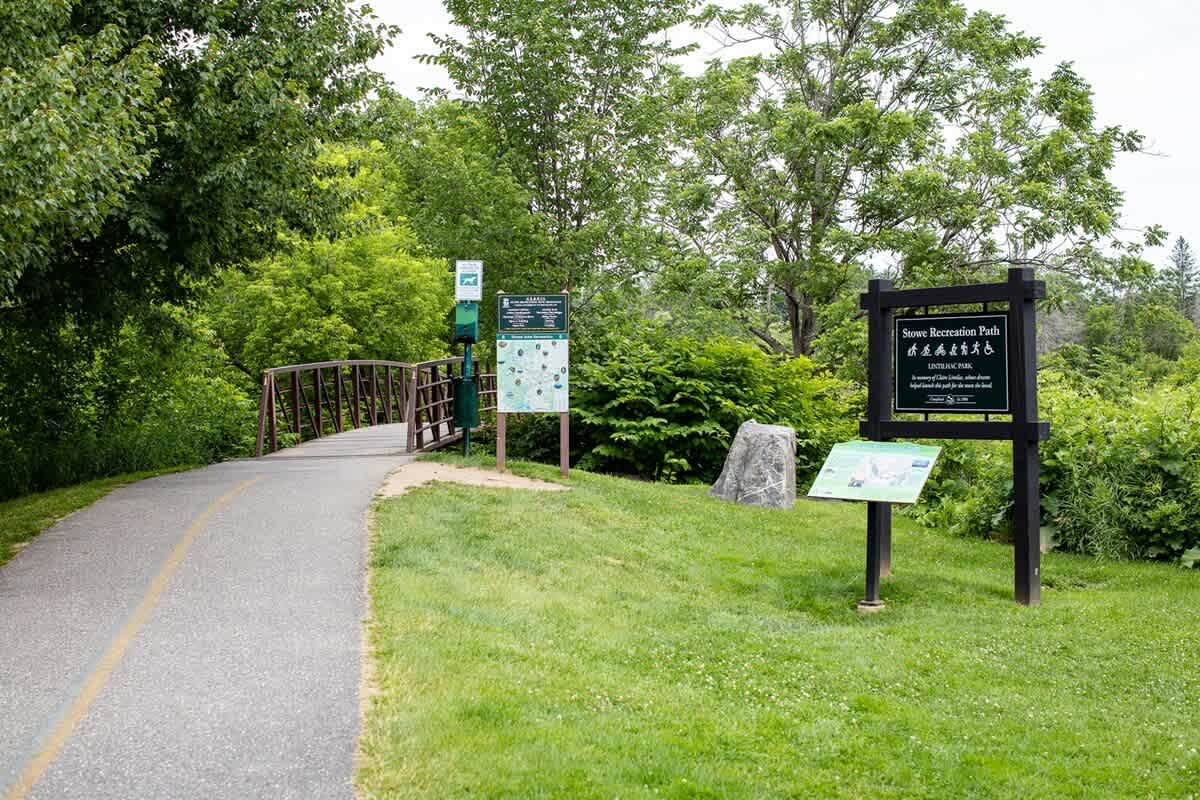 A paved recreation path curves towards a metal bridge, surrounded by lush green foliage and trees, with informational signs about the "Stowe Recreation Path" on the right.