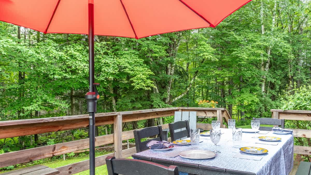 An outdoor dining table set for six with plates, glasses, and napkins is positioned under a large orange umbrella on a wooden deck, overlooking a vibrant green forest.