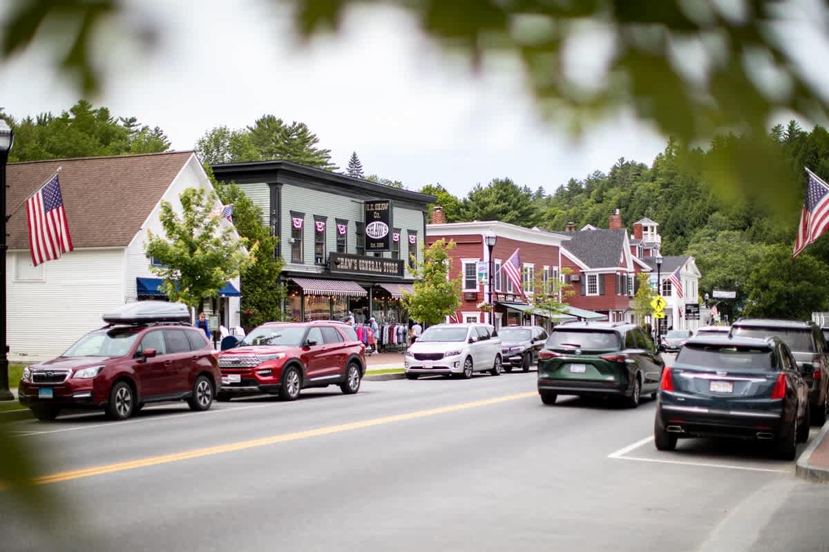 A charming street scene in a small town features cars parked along the roadside and historic buildings with American flags, surrounded by green trees under a cloudy sky.
