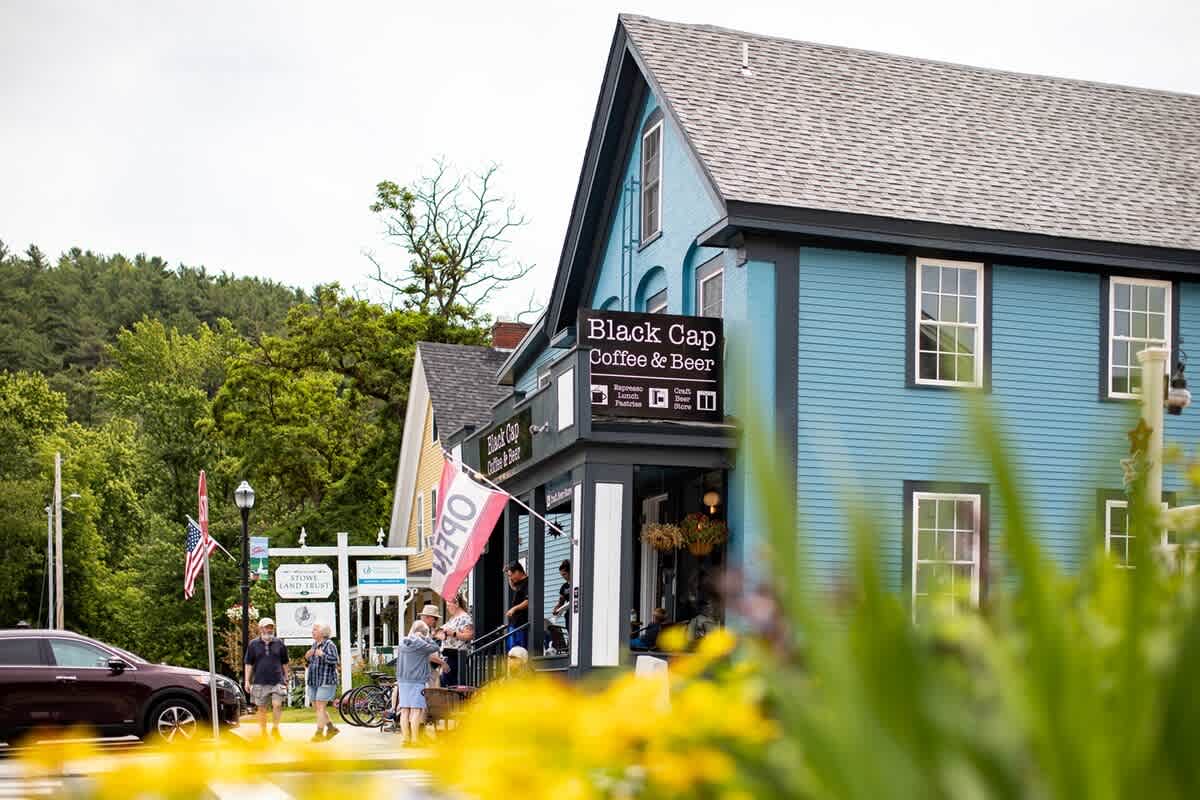 A vibrant blue building housing "Black Cap Coffee & Beer" stands prominently on a town street, with people gathered outside and lush green foliage in the foreground and background.