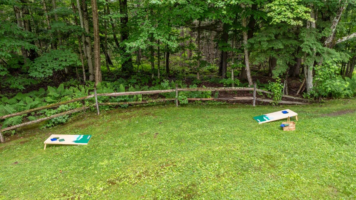 A spacious green lawn features two cornhole boards set up for a game, bordered by a rustic wooden fence and a dense forest in the background.
