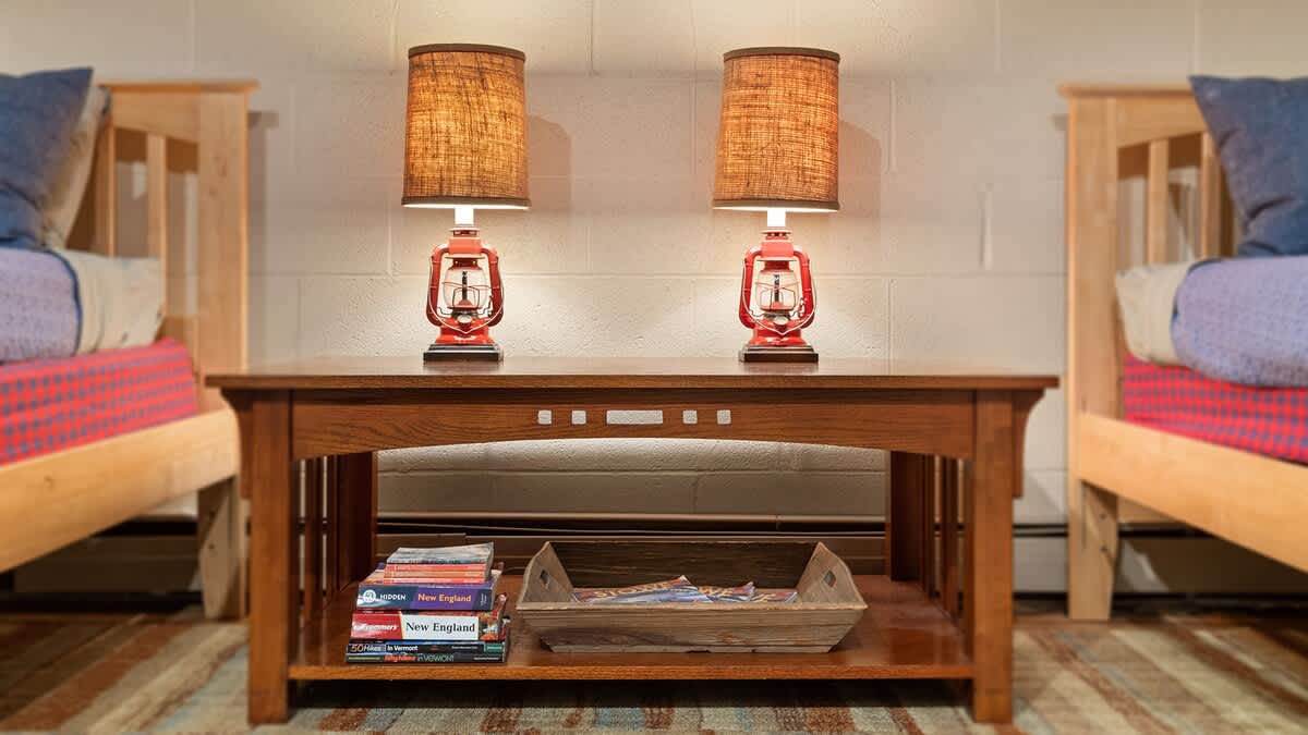 A close-up view of the wooden coffee table between two twin beds, showcasing two red lantern-style lamps with fabric shades. Books and a decorative tray are neatly arranged on the lower shelf.