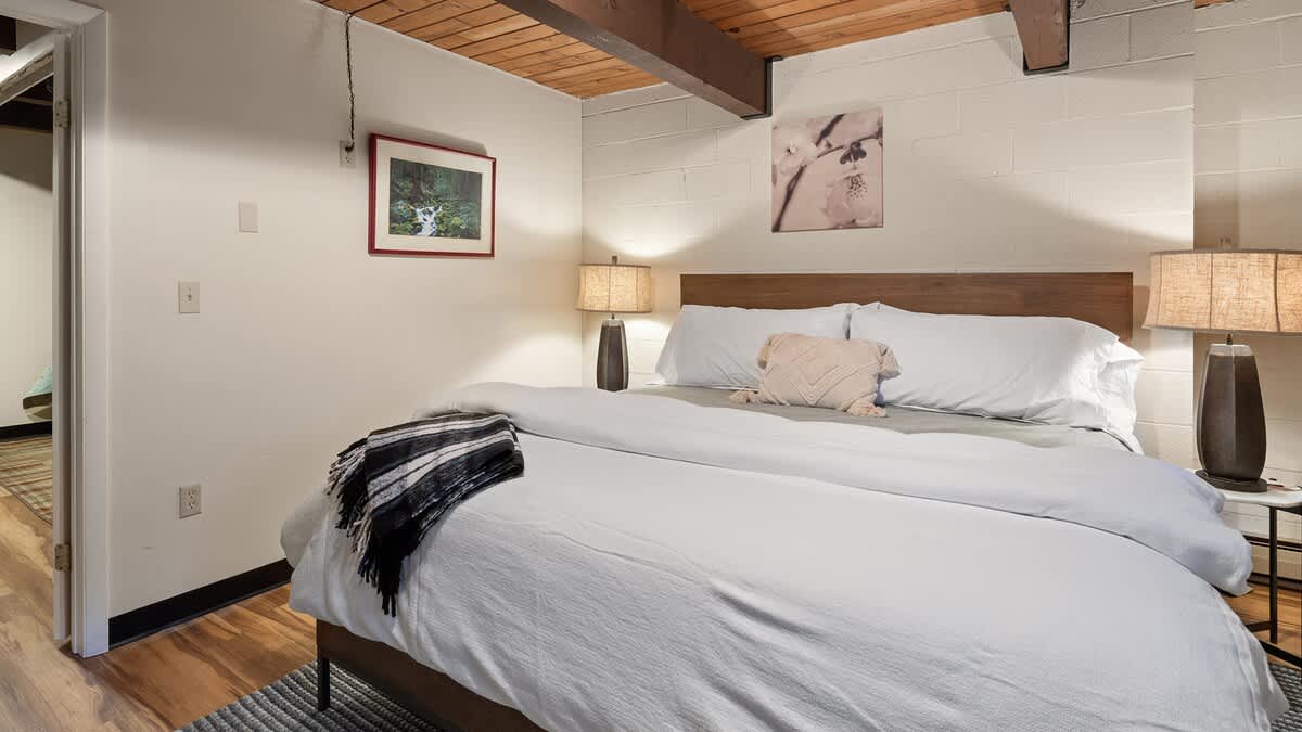 A bedroom with a wooden headboard bed, white bedding, and a striped throw blanket, set against white walls with exposed wooden ceiling beams. Two matching lamps flank the bed, and framed art hangs above.