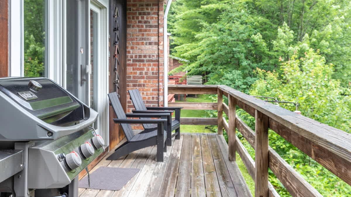 A gas BBQ grill sits on a wooden deck next to two dark Adirondack chairs, with a brick wall of the house on the left and a lush green forest visible beyond the wooden railing.