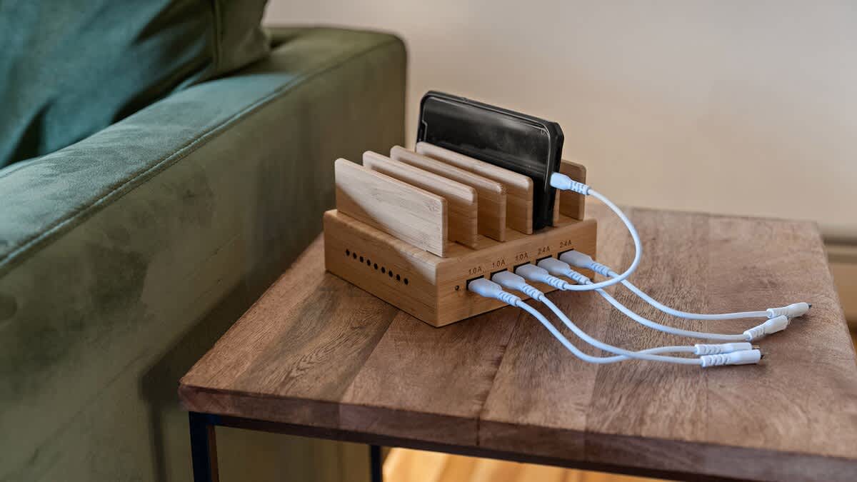 A wooden side table with a multi-device charging station, featuring a smartphone docked and multiple charging cables, positioned next to a green sofa.
