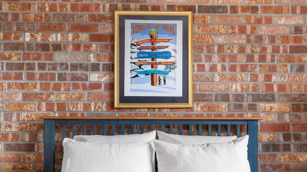 A bedroom featuring a blue wooden headboard against a prominent brick accent wall, with a framed "Vermont Ski Areas" poster hanging above the bed.