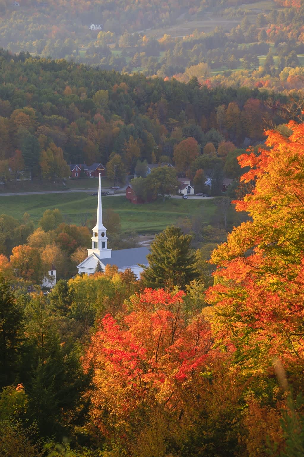 A breathtaking aerial view captures a picturesque village nestled in a valley, surrounded by a vibrant tapestry of autumn foliage in shades of orange, red, and green, with a white church steeple rising prominently. The scene highlights the stunning fall colors of the region.
