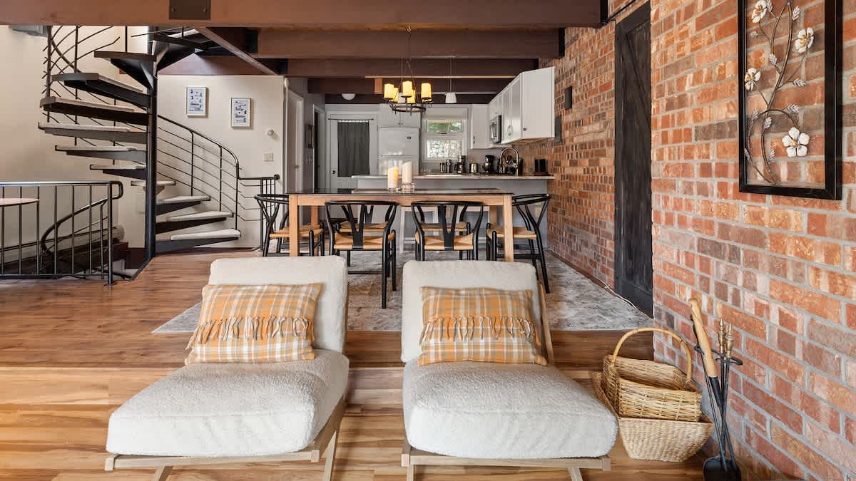 Spacious open-concept living area with two chaise lounges and a spiral staircase leading to an upper level, featuring exposed wooden beams, hardwood floors, and a dining area in the background.