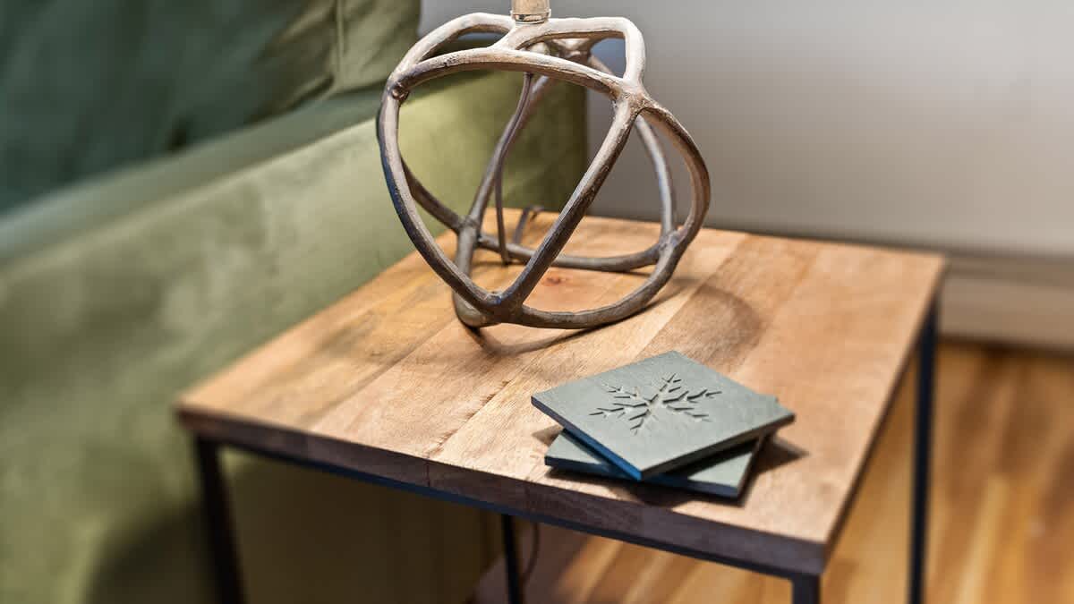 A wooden side table with a decorative, abstract metal sculpture and a stack of dark gray coasters with a snowflake design, positioned next to a green sofa.