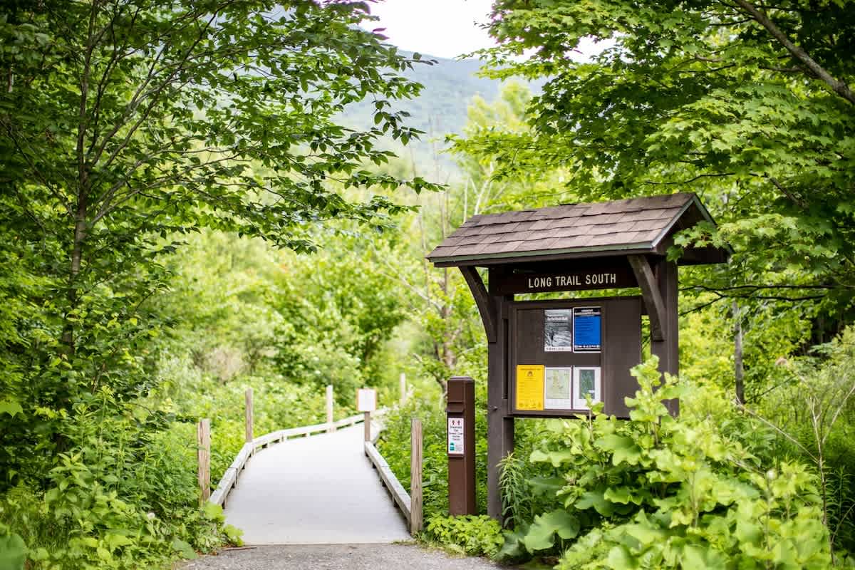A wooden boardwalk path winds into a vibrant green forest, flanked by dense foliage, with a prominent "Long Trail South" sign and an information board at the entrance. The setting suggests access to hiking and nature.