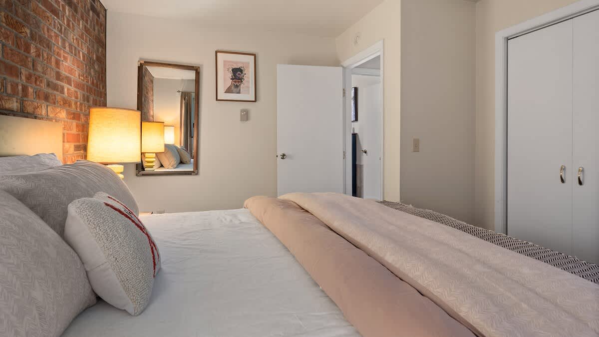 A cozy bedroom features a bed with white and light pink bedding, an exposed brick accent wall, and two bedside lamps casting a warm glow. A mirror and framed art adorn the light-colored walls.