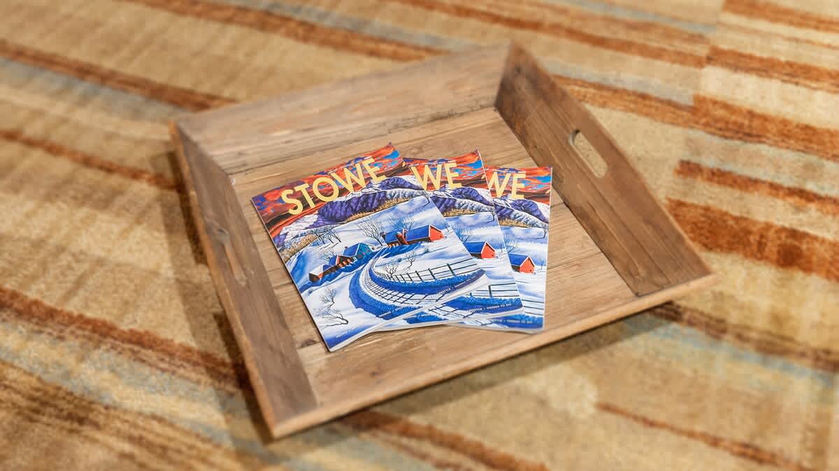 A close-up shot of three "Stowe" magazines resting on a rustic wooden tray, placed on a patterned rug or carpet. The magazines feature a winter landscape scene.