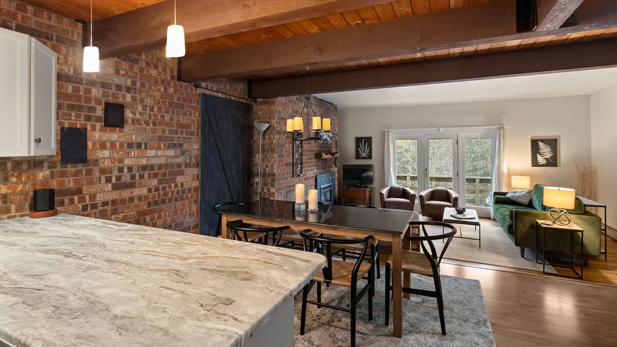 An open-concept living space showcasing a kitchen island with pendant lights, a dining area with a dark granite table and chairs, and a living room with a green sofa and fireplace, all against a brick wall and exposed wooden beams.