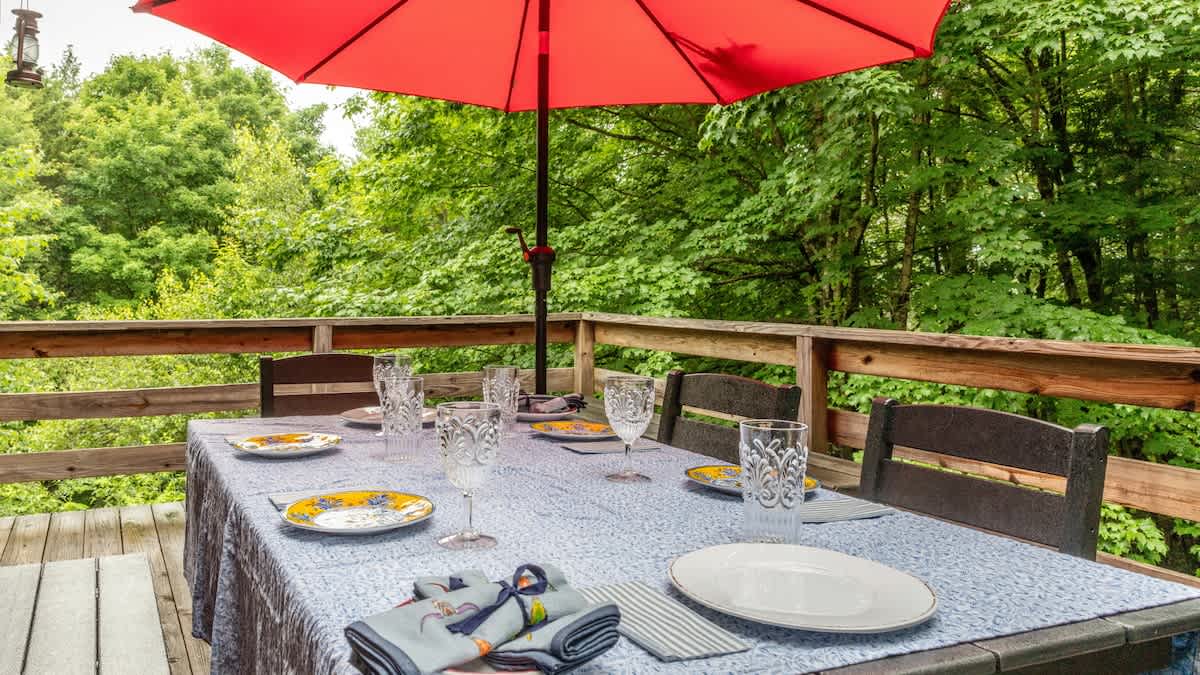 An outdoor dining table, set with plates, glasses, and napkins, is covered by a blue tablecloth and shaded by a large red umbrella on a wooden deck. Lush green trees surround the area.