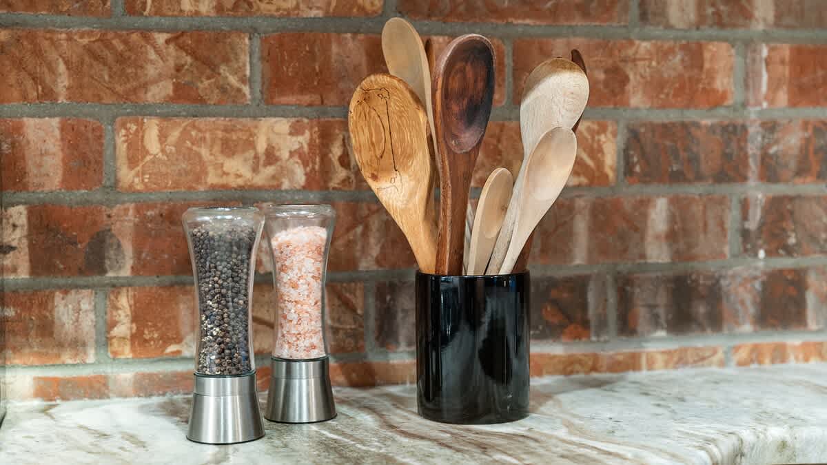 A close-up of a kitchen countertop with a brick backsplash, featuring two modern salt and pepper grinders and a black ceramic holder filled with various wooden cooking utensils.