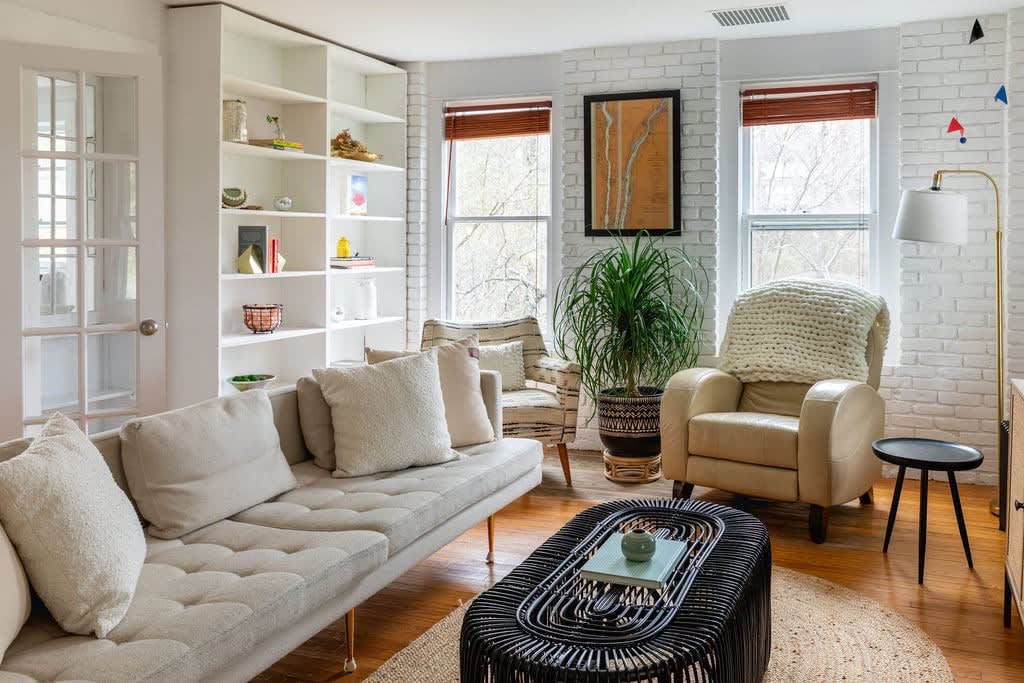 Inviting living room featuring a long light-colored sofa, built-in white shelving, and a black rattan coffee table. White painted brick walls and large windows with wooden blinds add to the bright and airy feel.