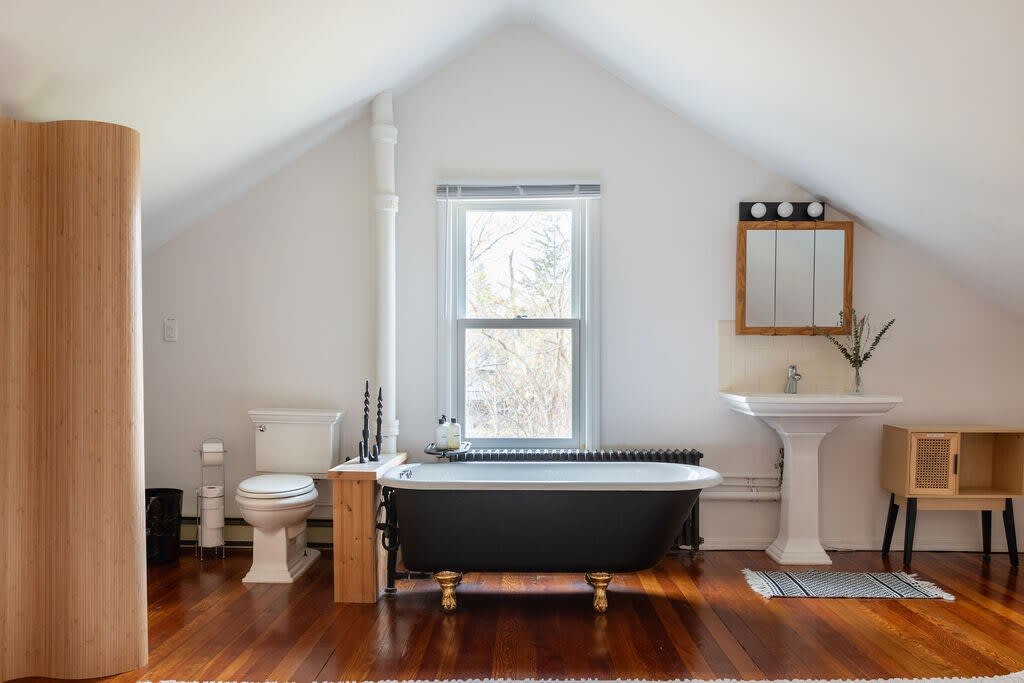A bright bathroom with a sloped ceiling features a classic black clawfoot tub with golden feet, a white pedestal sink, and a toilet. Hardwood floors and a large window complete the space.