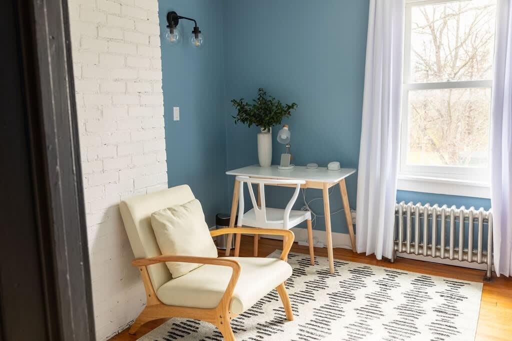A cozy office space with a striking blue accent wall, white brick fireplace, a light wood desk with a white chair, and a cream armchair on a geometric patterned rug.