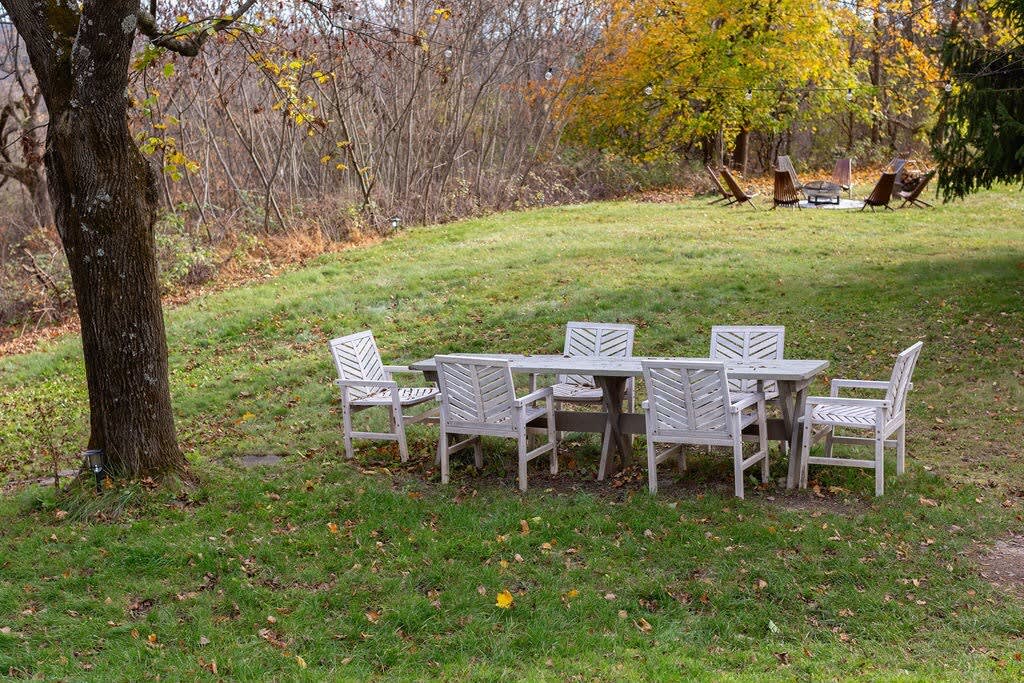 A white outdoor dining table with six matching chairs is placed on a green grassy lawn, surrounded by trees with autumn foliage in the background.