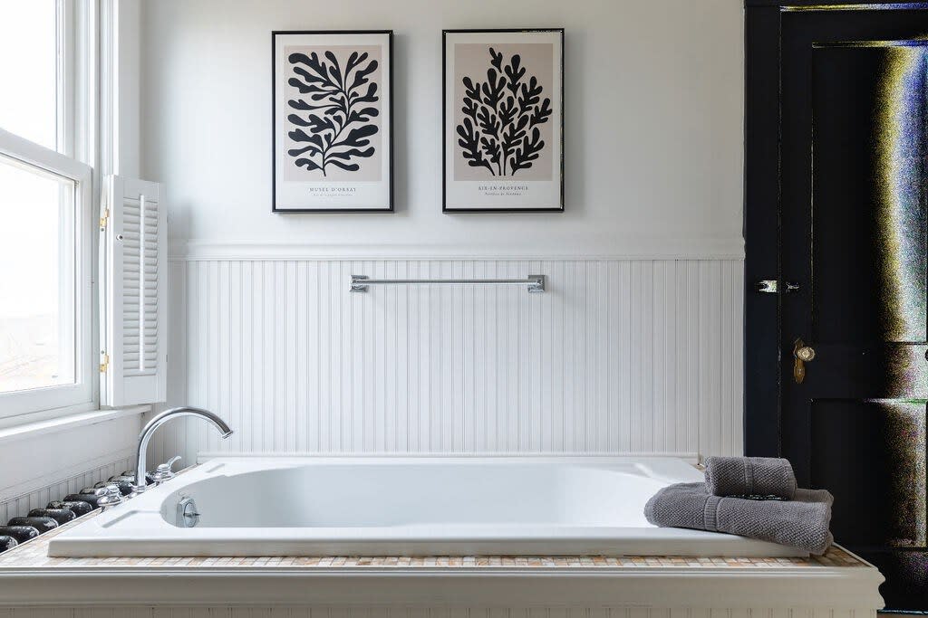 A clean bathroom featuring a built-in bathtub with a tiled surround, white beadboard wainscoting, and two framed botanical prints on the wall above. A dark door with a vintage doorknob is visible on the right.