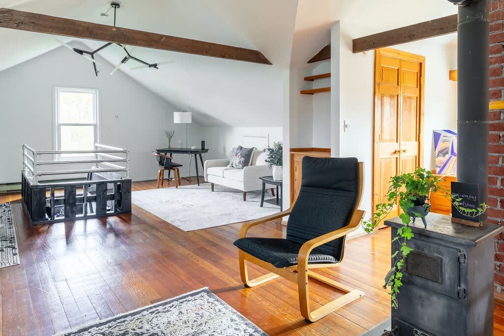 A spacious living area with sloped ceilings and exposed wooden beams features a black wood-burning stove, a light-colored sofa, and a desk area. Hardwood floors are partially covered by a light patterned rug.