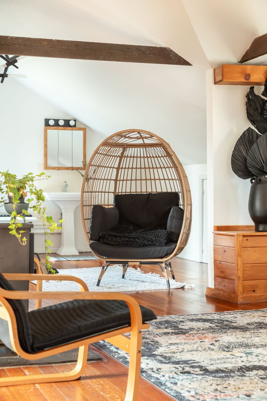 A comfortable seating nook features a large wicker egg chair with black cushions and a modern wooden lounge chair, set on hardwood floors. The bathroom with a pedestal sink is visible in the background.