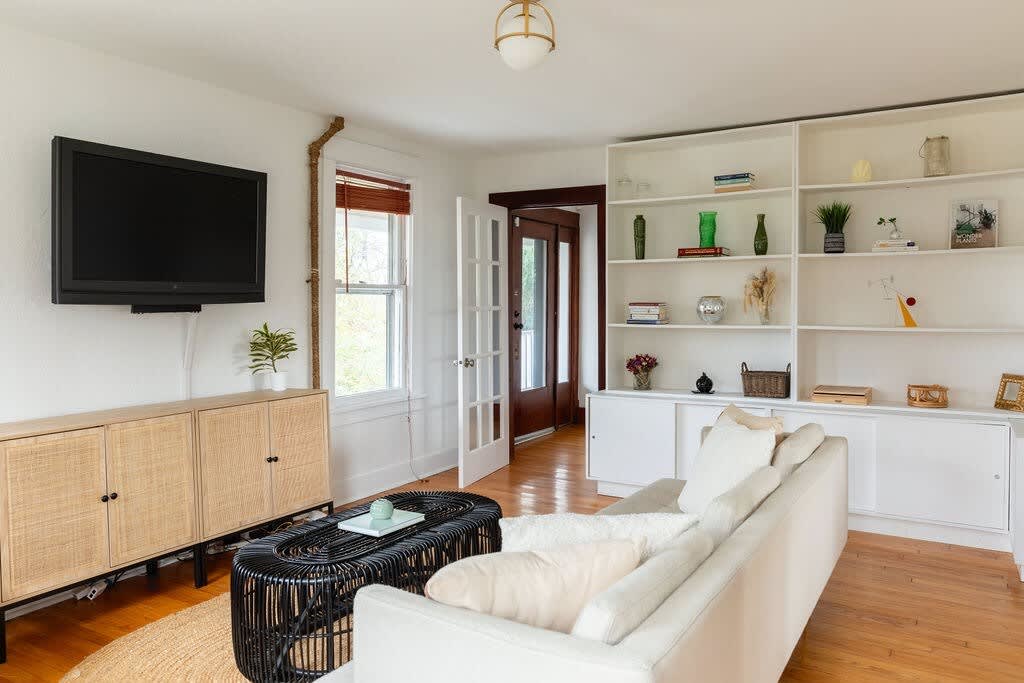 Another view of a living area with a light-colored sofa, a wall-mounted TV above a rattan cabinet, and built-in white shelving. French doors lead to another room, and hardwood floors run throughout.