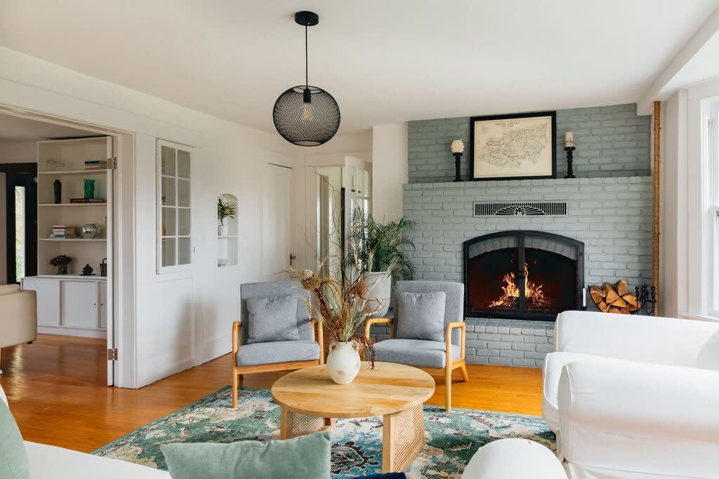 A cozy living room features a light gray brick fireplace with a burning fire, flanked by two modern gray armchairs and a light wood coffee table on a patterned green rug. A white sofa is partially visible, and a black mesh pendant light hangs from the ceiling.