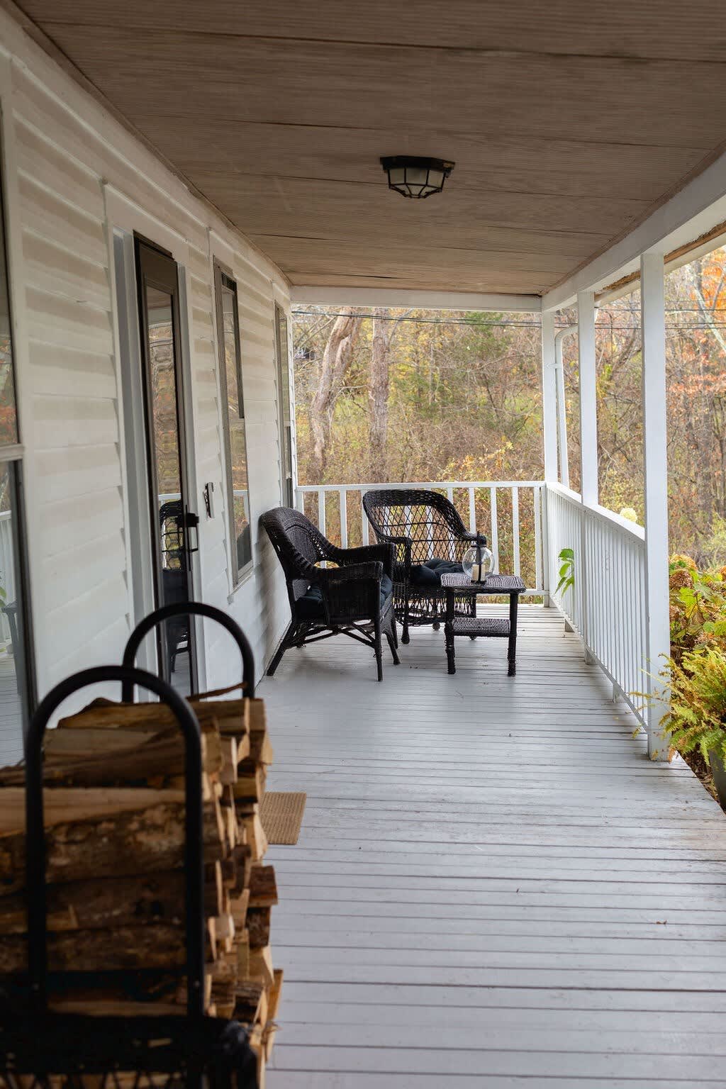 A covered porch features two dark wicker chairs and a small table, with a metal rack holding stacked firewood in the foreground, overlooking a wooded area.