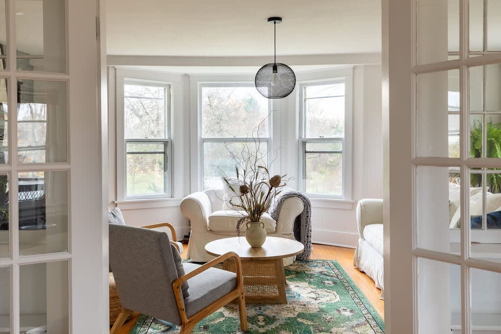 A view into a sunlit living room through white French doors, showcasing a bay window area with white armchairs and a round wooden coffee table. A green patterned rug covers the hardwood floor, and a black mesh pendant light hangs centrally.