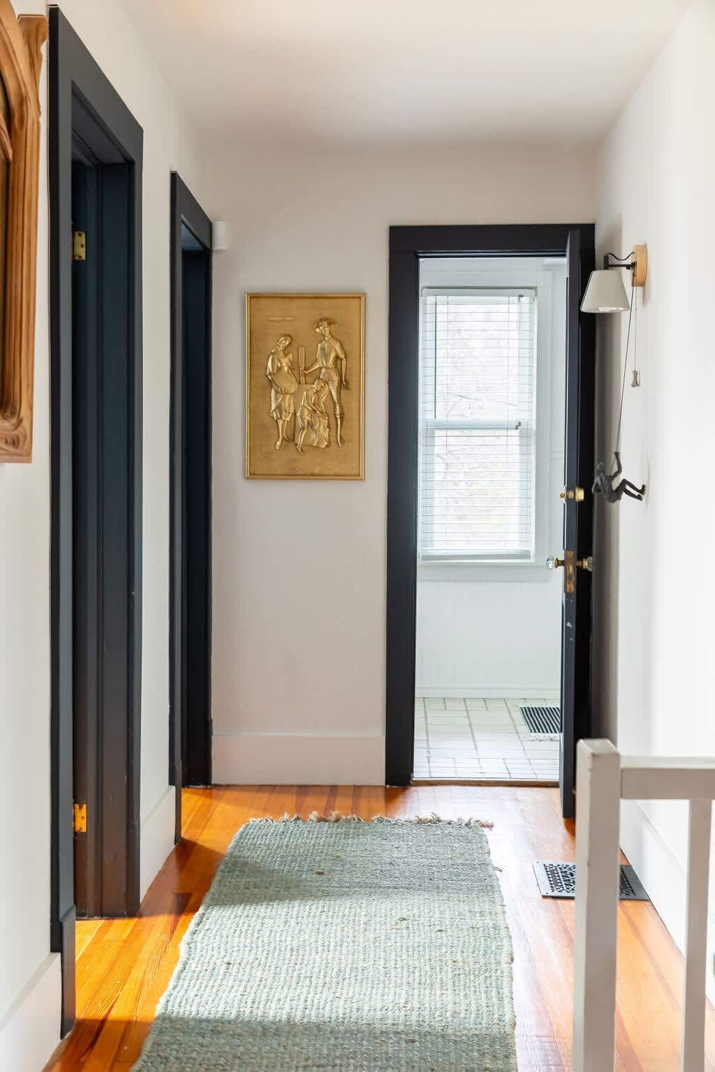 A narrow hallway with polished hardwood floors, white walls, and dark trim around doorways, featuring a light green textured runner rug and a gold framed relief artwork.