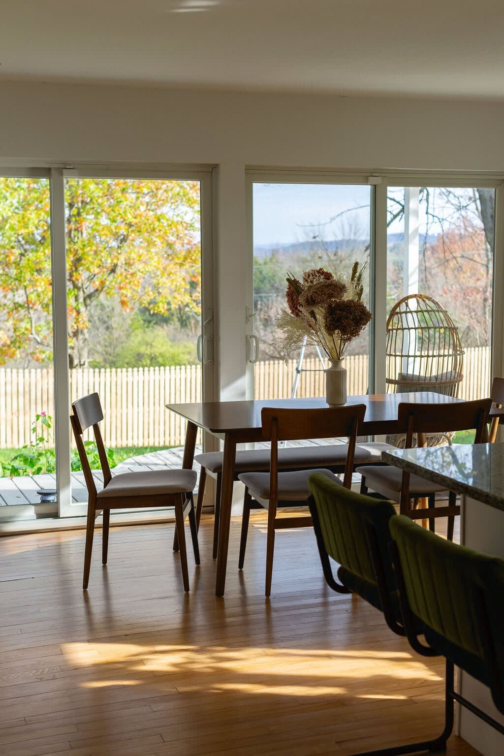 A dining area with a dark wood rectangular table and matching chairs, set against large sliding glass doors that open to a deck and a fenced backyard with distant hills. A vase with dried flowers sits on the table, and hardwood floors extend throughout.