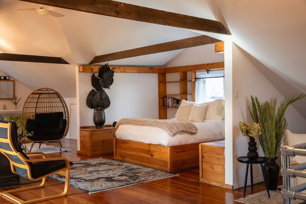 A cozy attic bedroom features a wooden platform bed with white bedding and a chunky knit throw, exposed wooden beams, and a wicker egg chair with black cushions. A patterned rug covers the hardwood floor.