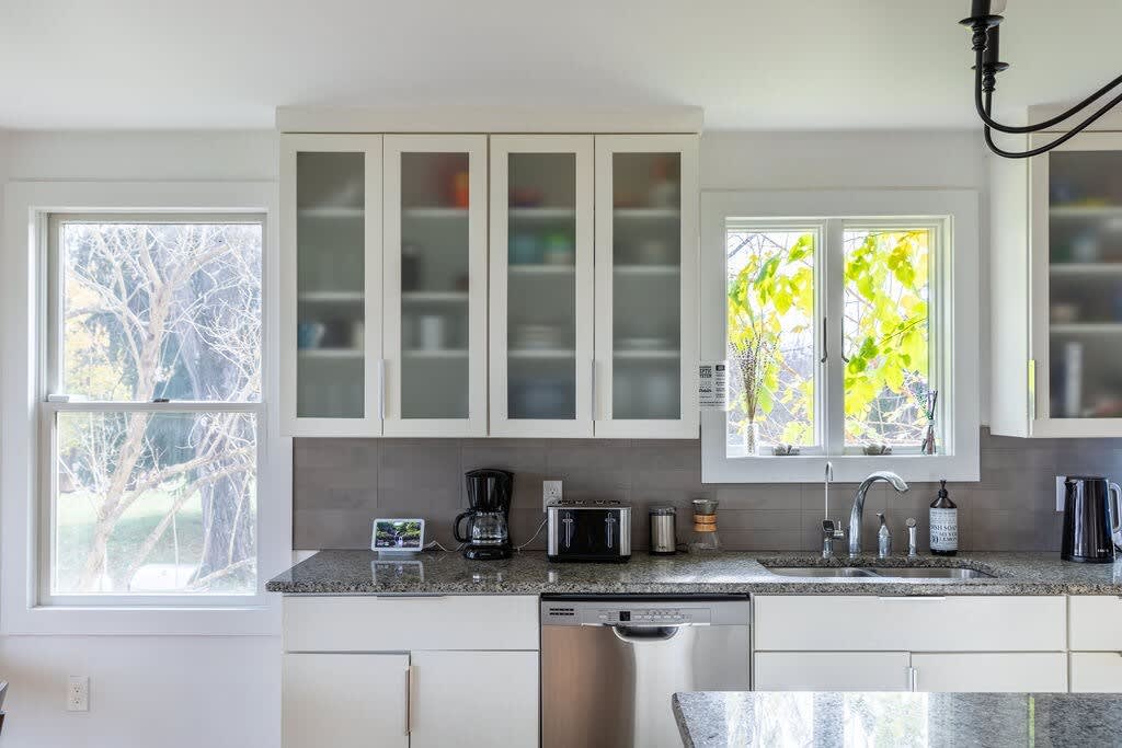 A close-up of the kitchen counter area shows white cabinets with frosted glass doors, granite countertops, and a stainless steel sink with a modern faucet. Various small appliances like a coffee maker and toaster are visible, with windows offering natural light.