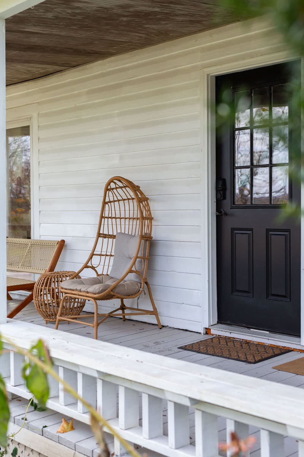 A tall, natural wicker egg chair with a light cushion is positioned on a white wooden porch, beside a black front door with glass panes.