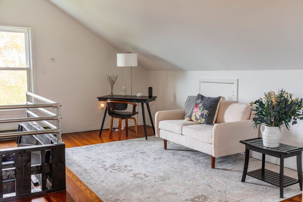 A bright living space with sloped ceilings includes a small light-colored sofa with decorative pillows, a black desk with a chair, and a large window. A light patterned rug defines the seating area on hardwood floors.