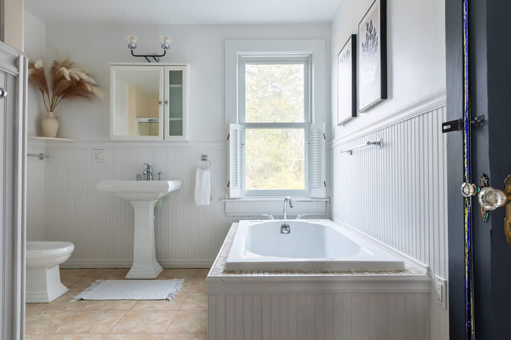 A spacious white bathroom with a pedestal sink, toilet, and a built-in bathtub, all surrounded by white beadboard wainscoting and light-colored floor tiles. A window with white shutters provides natural light.