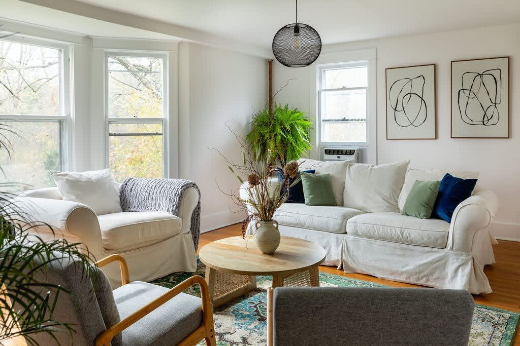 A bright living room with white slipcovered sofas and armchairs, accented with green and blue throw pillows, surrounds a round wooden coffee table. Large windows overlook a lush outdoor scene, and abstract art hangs on the white wall.